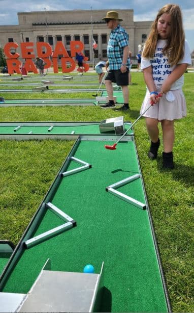 A child playing mini golf on a bright green course at an outdoor event, with other participants and a ‘Cedar Rapids’ sign visible in the background.