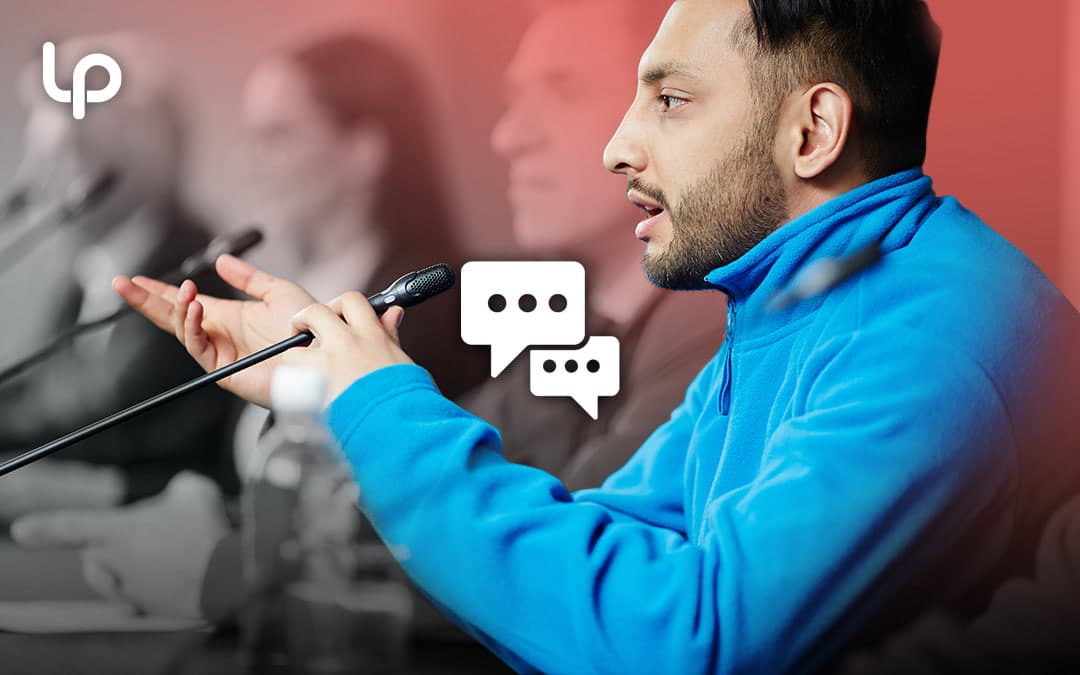 A man in a blue fleece jacket speaks into a microphone at a press conference or panel discussion, with a chat bubble icon overlay and the Lukas Partners logo in the corner.